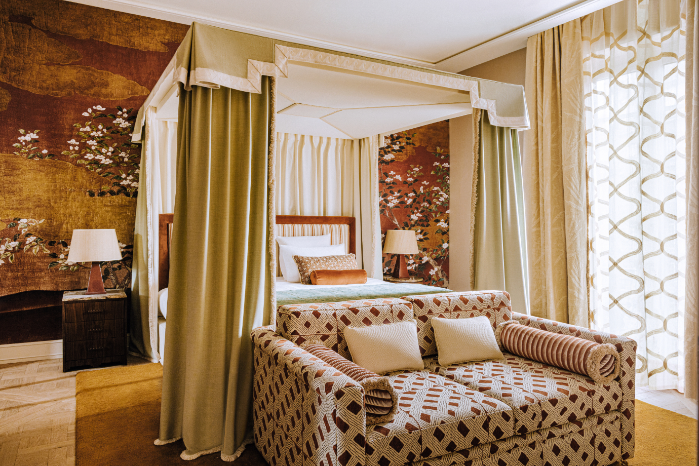 A deluxe room at Saint James Paris featuring a pea green canopy bed, spice-toned velvet pillows, mid-century modern lamps, rust-colored silk chinoiserie wallpaper, a burgundy and cream patterned loveseat and floor to ceiling windows