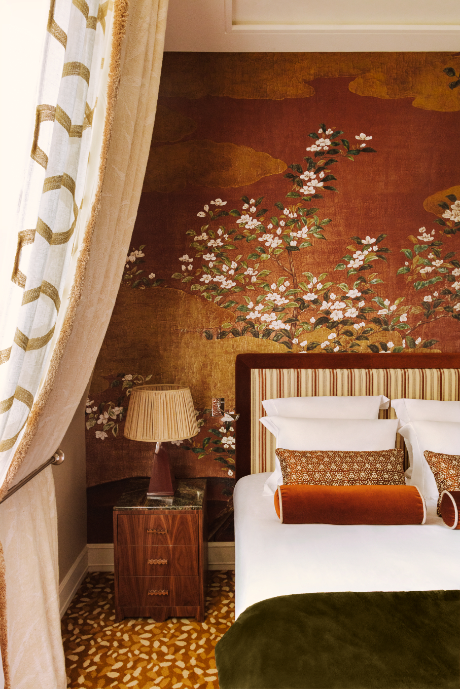 A cinnamon colored room at Saint James Paris with maximalist interiors, Asian-inspired chinoiserie wallpaper, striped headboards, raffia lampshades, velvet pillows and thick beige curtains