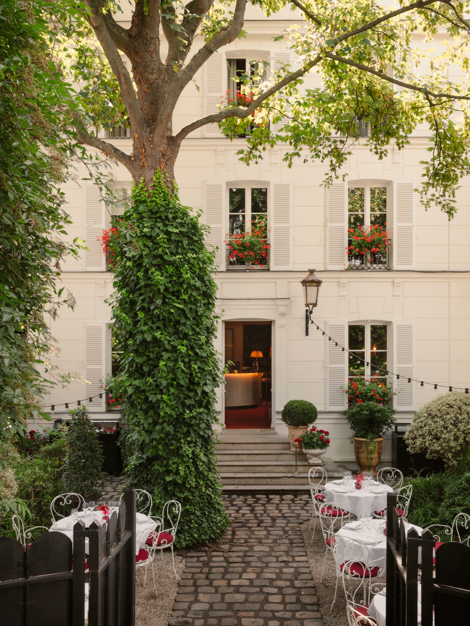 The hidden courtyard garden entrance of Hôtel Particulier Montmartre with crisp white linens + French iron garden chairs in front of a white 19th townhouse with red geraniums filling the shuttered balconies