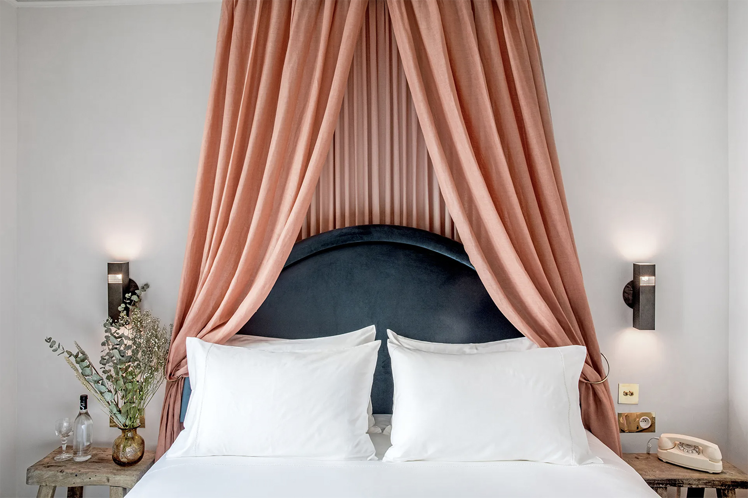A dramatic pink linen canopy bed with a navy velvet headboard and vintage olive wood stools serving as side tables at Hotel des Grands Boulevards