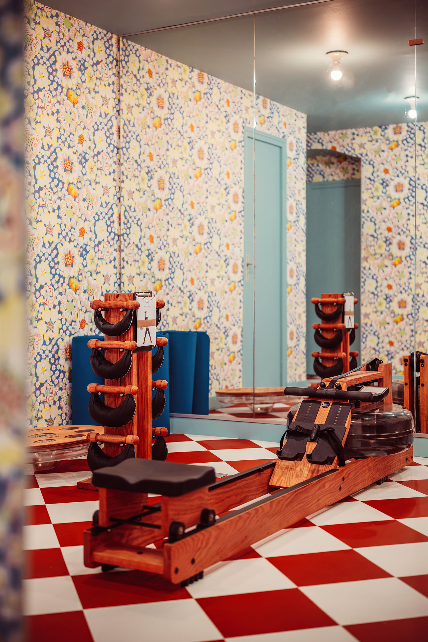 The playfully designed gym room at Hotel les Deux Gares with a vintage rowing machine, red and white checkerboard floors and cartoonish blue, purple and yellow floral walls