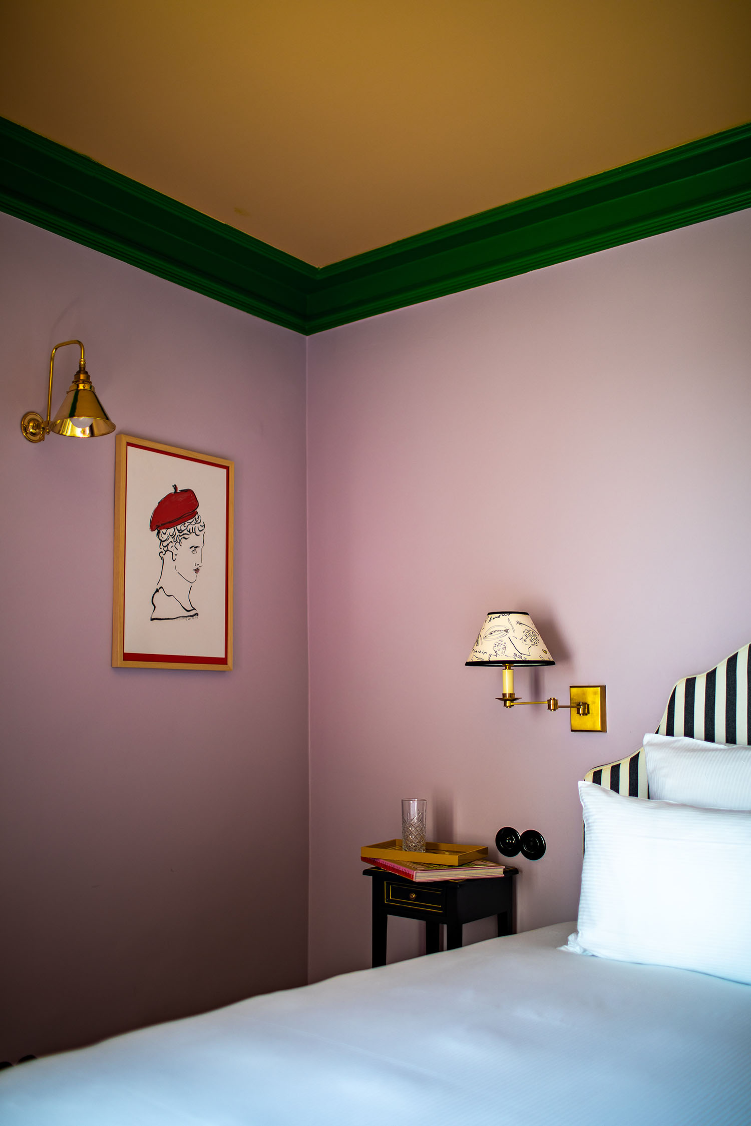 An lavender guest room at Hotel les Deux Gares with a green + white striped headboard, mural-sketched wall lamp shades, and a Greco-Roman inspired wall illustration featuring a red beret