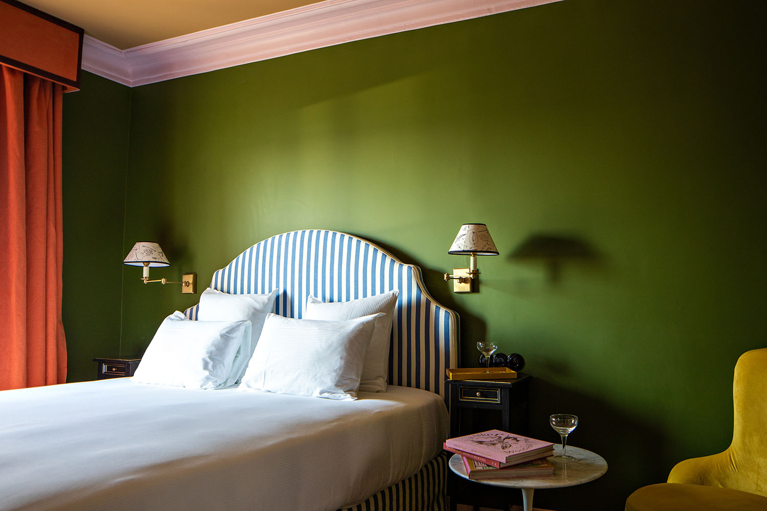 An olive green guest room at Hotel les Deux Gares with a blue and white striped headboard, mural-sketched wall lamp shades, a mustard velvet arm chair and a small white marble side table