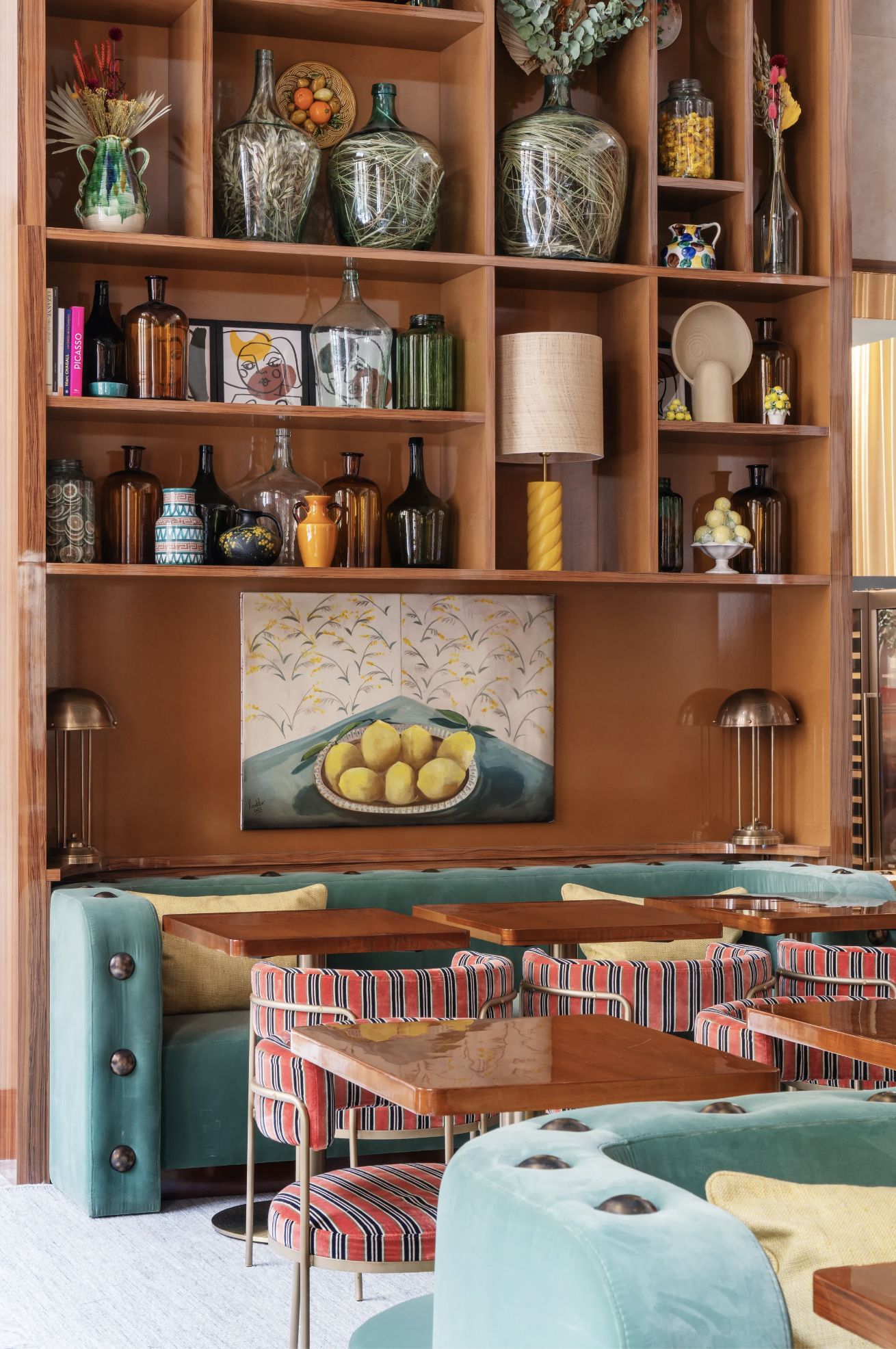 Eclectic interiors of Mimosa Paris iwth teal velvet banquette seating, striped fabric chairs and a wooden bookshelf filled with mismatched glass jugs and urns